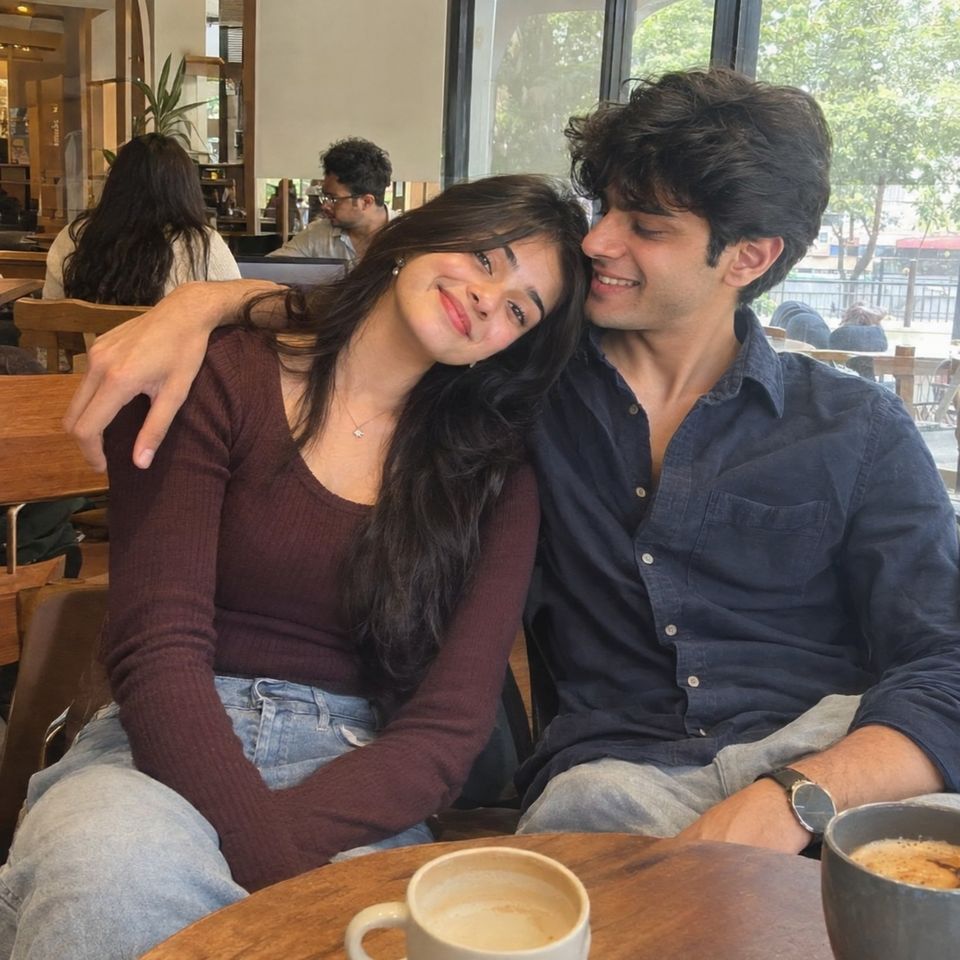 A couple at a café table, her head resting on his shoulder, two coffees in front of them